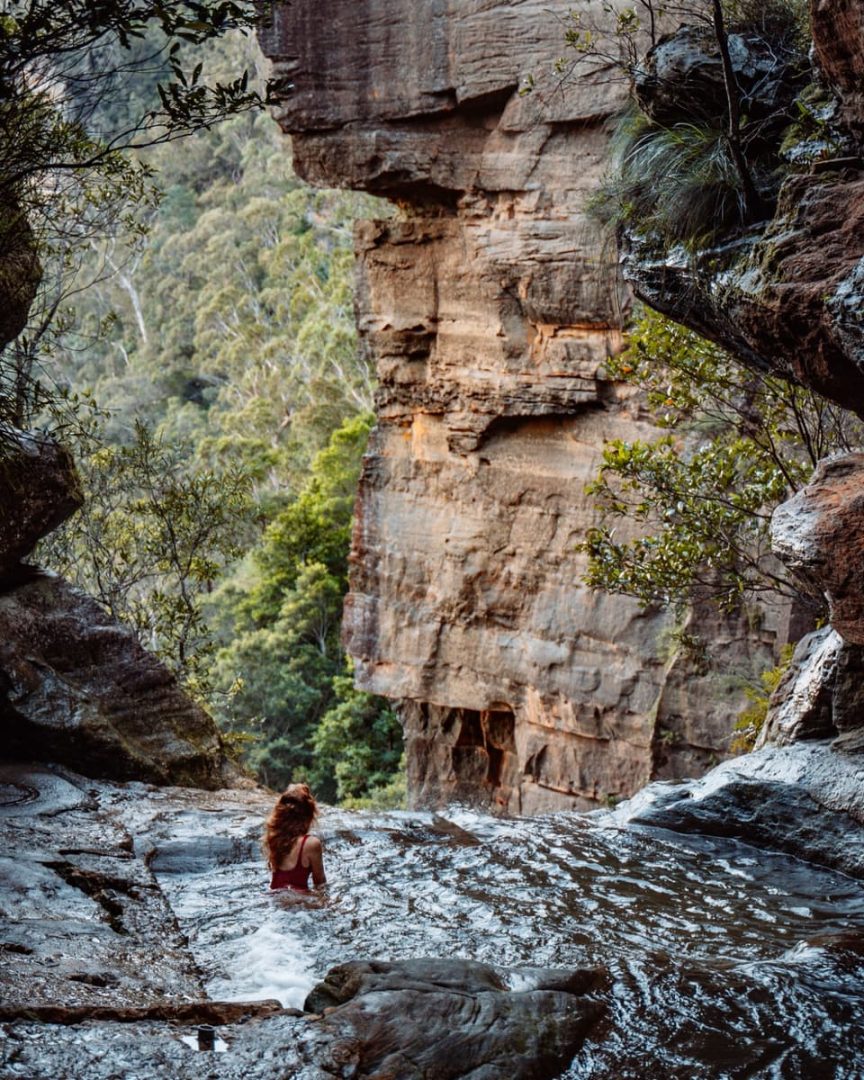 Woman sitting in a natural secret infinity pool in the Blue Mountains with water flowing around her, facing a dramatic sandstone cliff and lush forest below.