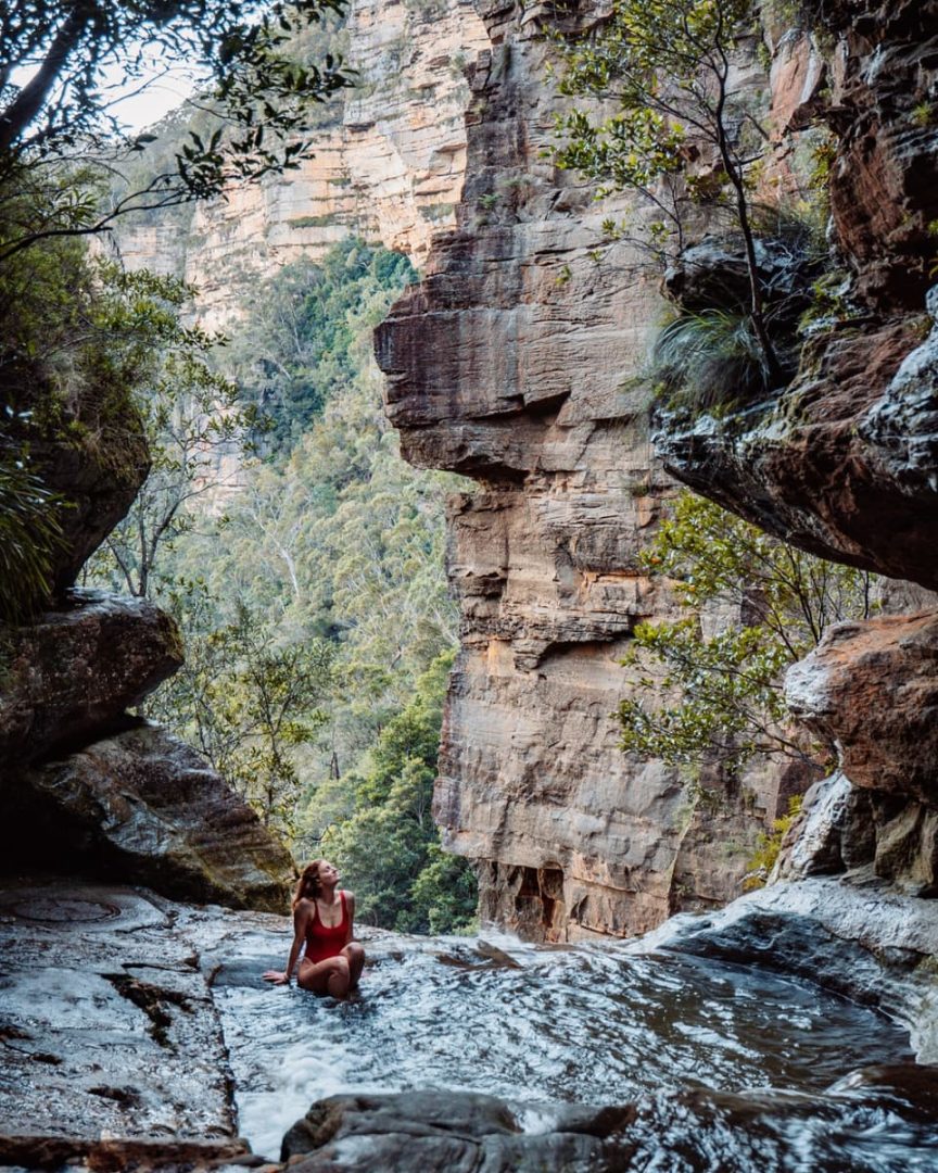 Woman in red swimsuit sitting in the secret infinity pool Blue Mountains, gazing up at the cliffs above as water flows gently past her.