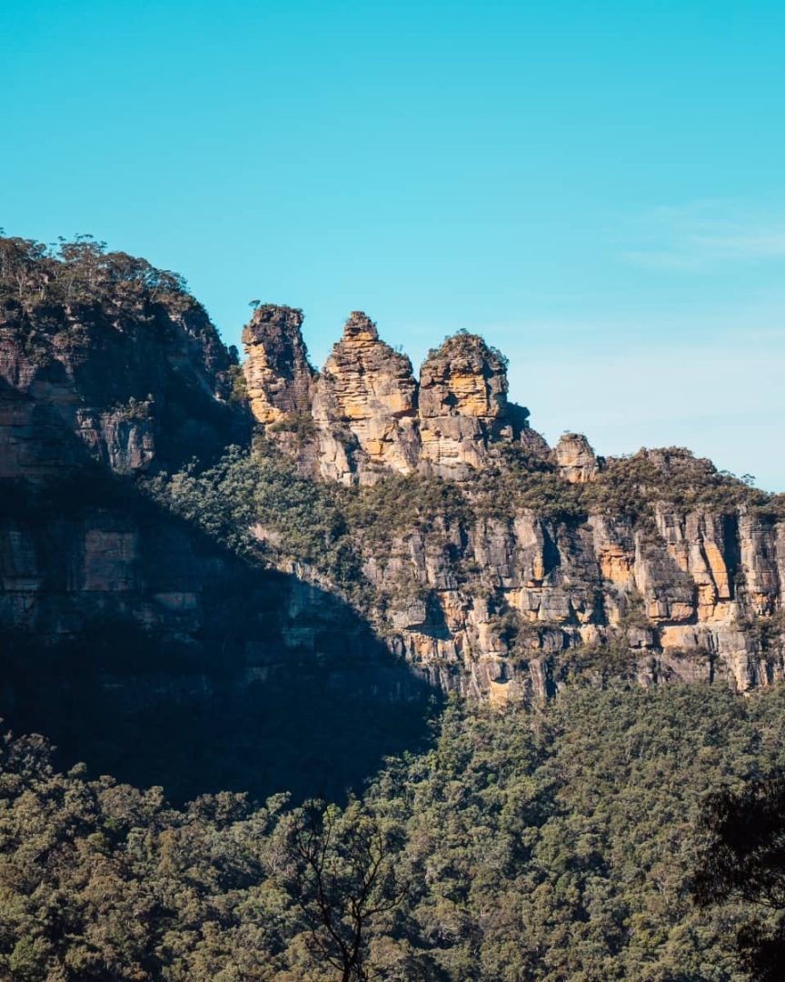 Distant view of the Three Sisters and surrounding cliffs of the Blue Mountains in NSW, with green forest below and a soft blue sky above.