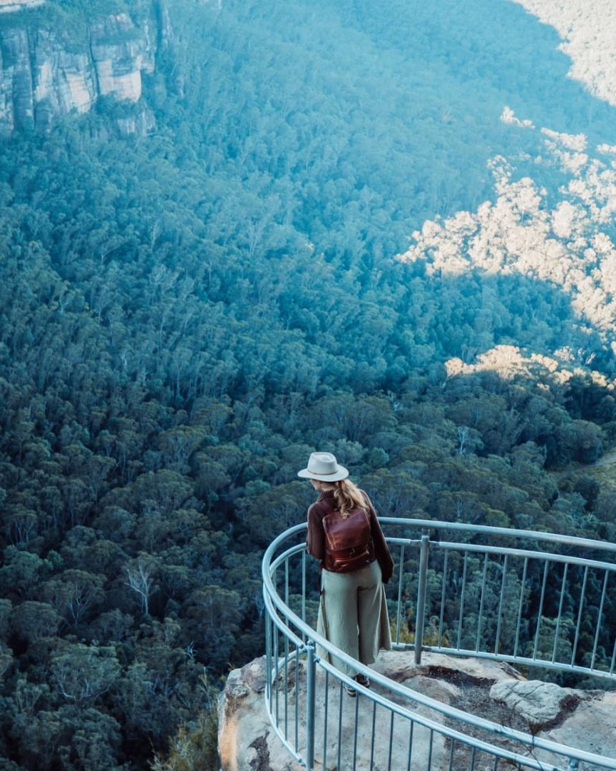 Woman wearing a hat and backpack looking out from a fenced lookout over the dense eucalyptus forest in the Blue Mountains valley below.
