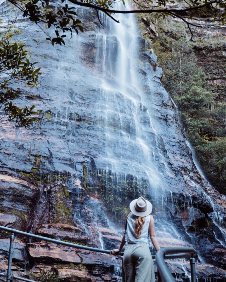 Woman with a white hat and ponytail standing at a railing, admiring the Bridal Veil Falls in the Blue Mountains, surrounded by mossy rock face. It's the halfway point to the secret infinity pool in Leura Cascades