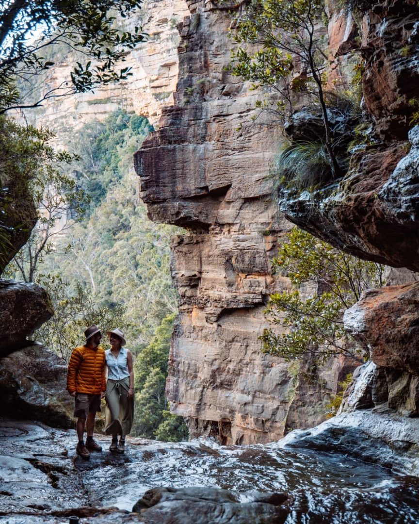 Chris and Bec standing on wet rocks above the secret infinity pool Blue Mountains, surrounded by towering cliffs and lush bushland in soft afternoon light.