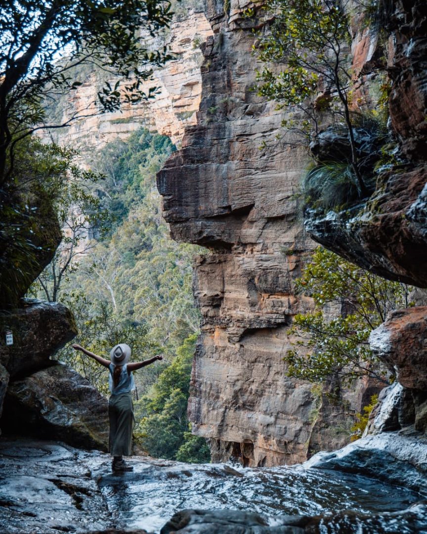 Woman standing with arms raised beside the secret infinity pool in Leura Cascades, Blue Mountains, facing dramatic cliff walls and forest valley below.