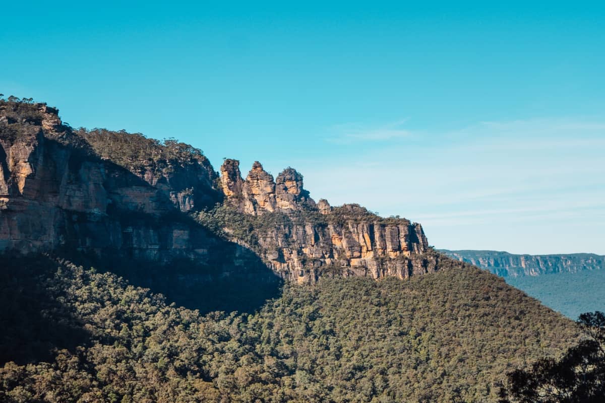 Distant view of the Three Sisters and surrounding cliffs of the Blue Mountains, with green forest below and a soft blue sky above.
