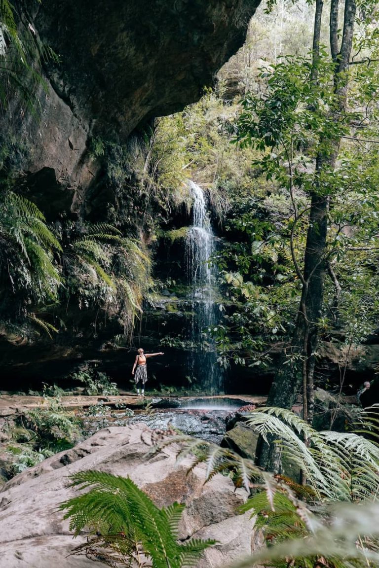 A woman stands on a rock pointing toward a narrow waterfall cascading down a mossy cliff surrounded by dense forest along the Grand Canyon Track in Blue Mountains. The scene highlights the lush greenery and tranquil atmosphere of this popular hiking spot.