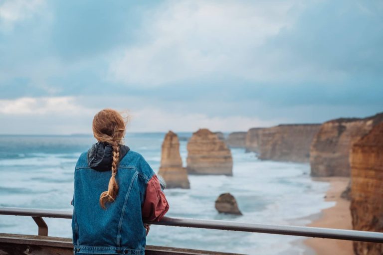 A woman in a denim jacket with a long braid looks out over the iconic limestone stacks of the Twelve Apostles along the southern coast, a must-see stop on any great ocean road itinerary.