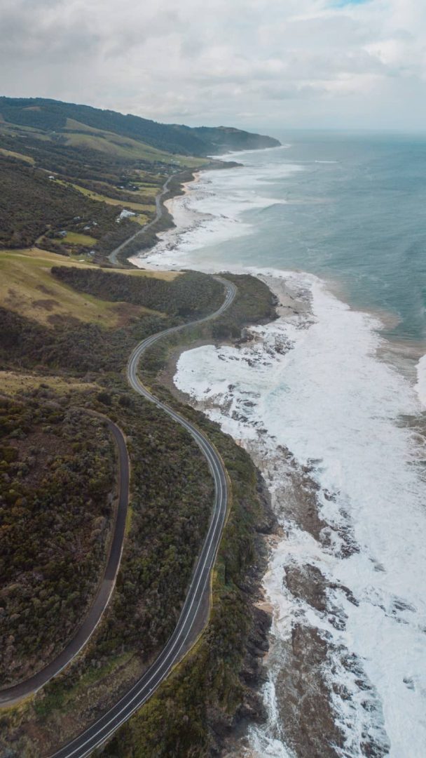 A winding stretch of road hugs the rugged coastline with cliffs and waves below, capturing the dramatic seascapes featured in a scenic great ocean road itinerary.