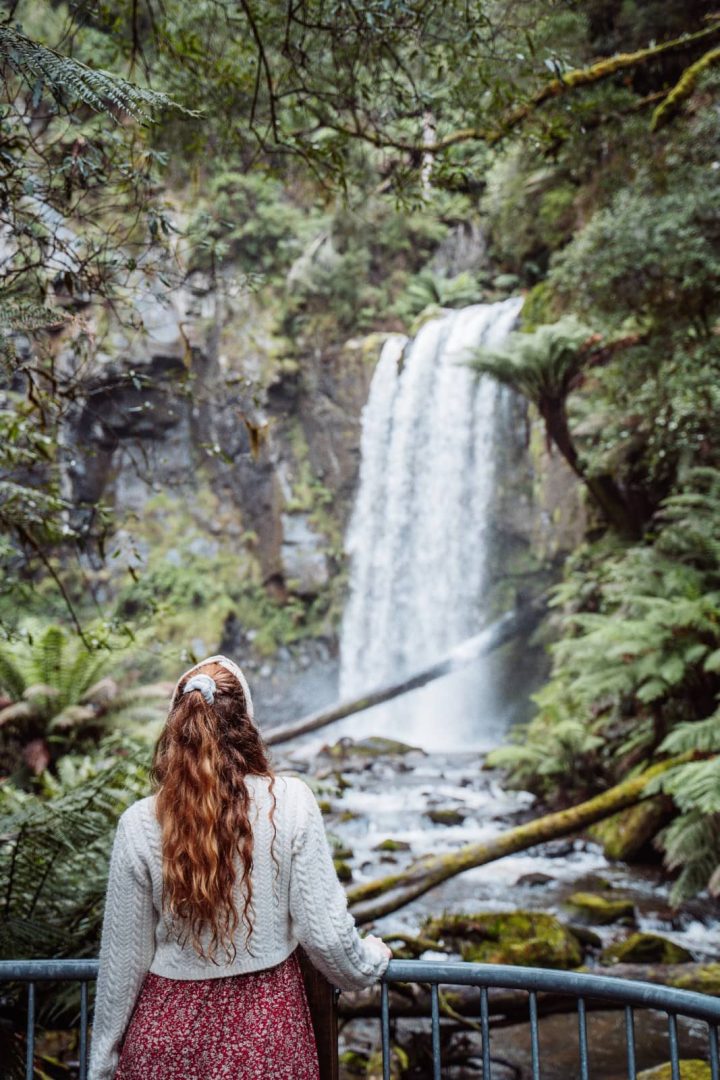 A woman in a floral skirt and jumper stands at a railing facing a tall forest waterfall, a tranquil moment on a great ocean road itinerary through the Otways.