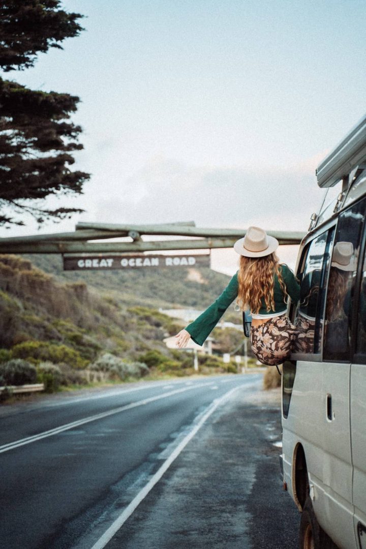 A woman leans joyfully out the window of a campervan below the “Great Ocean Road” sign, marking the start of one of Australia’s most popular road trip itineraries.