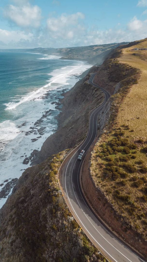 A campervan drives along a sharp coastal bend high above the ocean, showcasing the cliffside roads featured in an adventurous great ocean road itinerary.