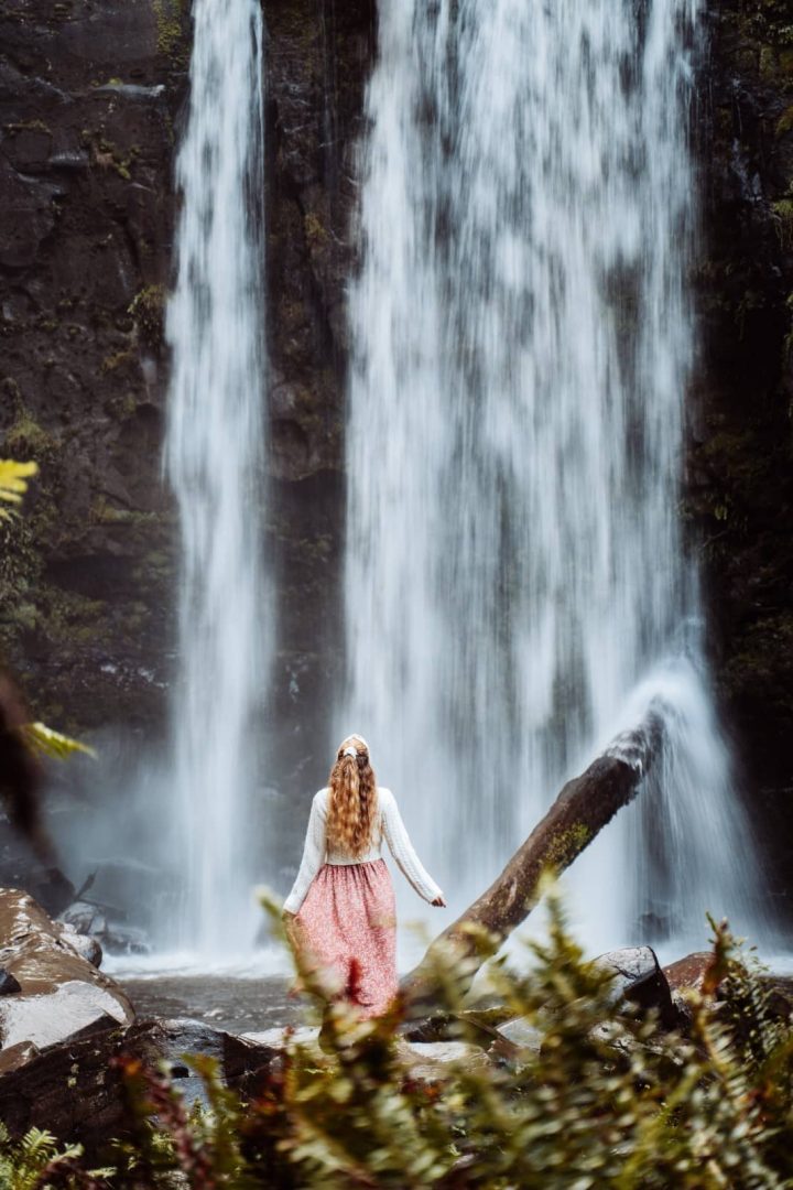 A woman in a pink skirt and white jumper stands in front of a tall, flowing waterfall surrounded by mossy rocks, a serene stop on many great ocean road itinerary routes.