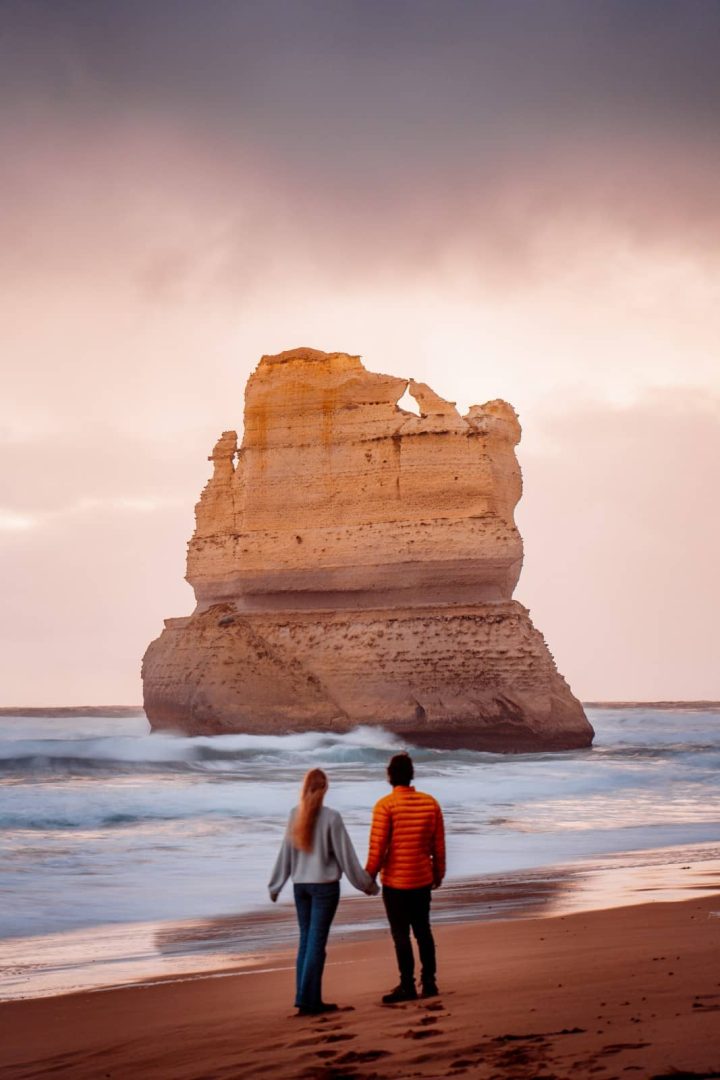 A couple holds hands while walking on the beach toward a large limestone sea stack during a glowing sunset, a romantic moment on a great ocean road itinerary.