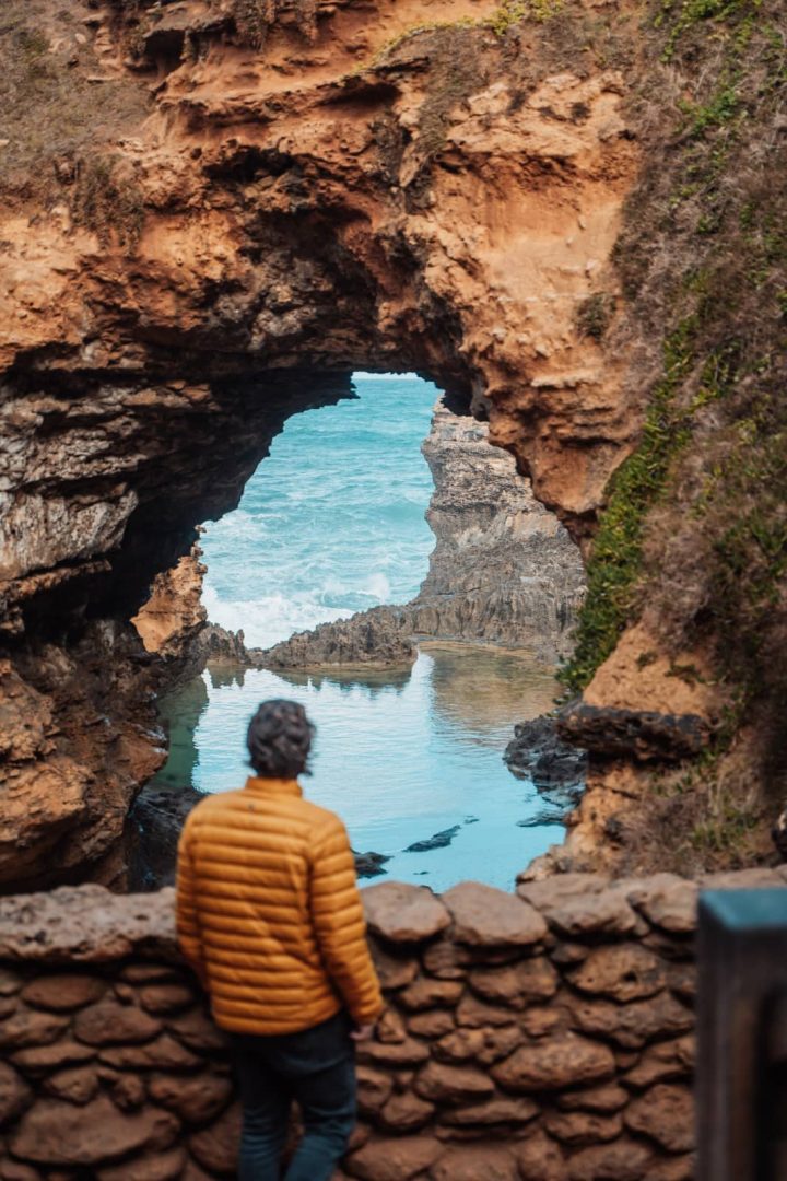 A man in an orange jacket stands before a natural rock arch with ocean visible through it, one of the geological wonders along the great ocean road itinerary.