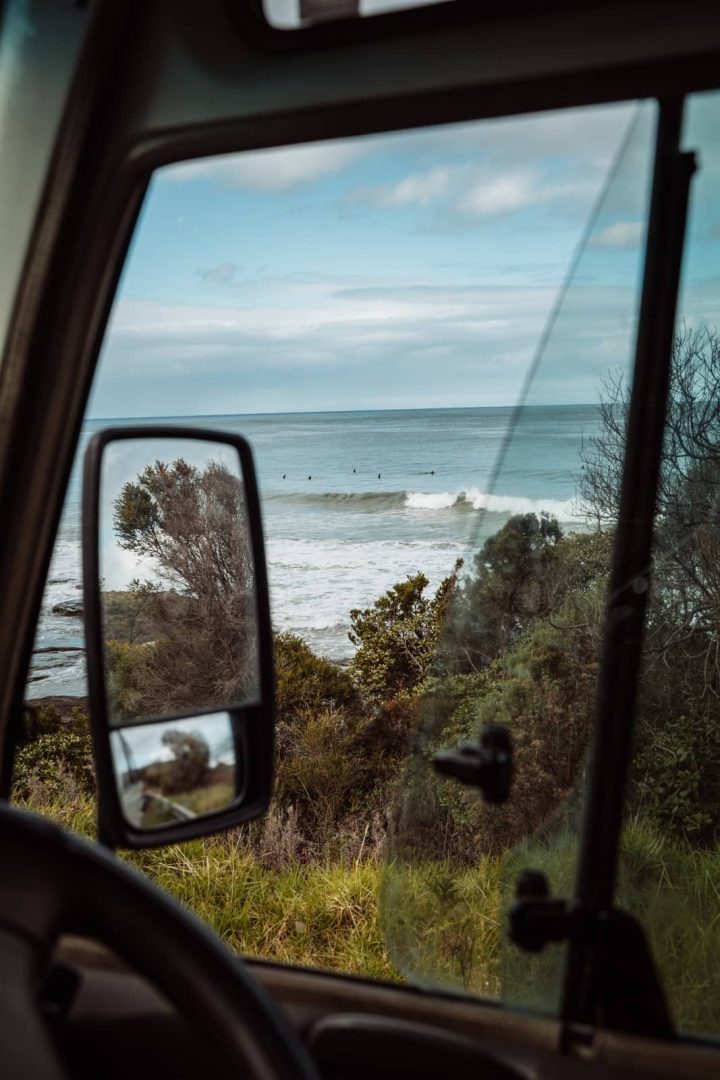 Ocean waves are seen through the rear view window of a parked campervan, showing the laid-back beach vibes along a great ocean road itinerary.