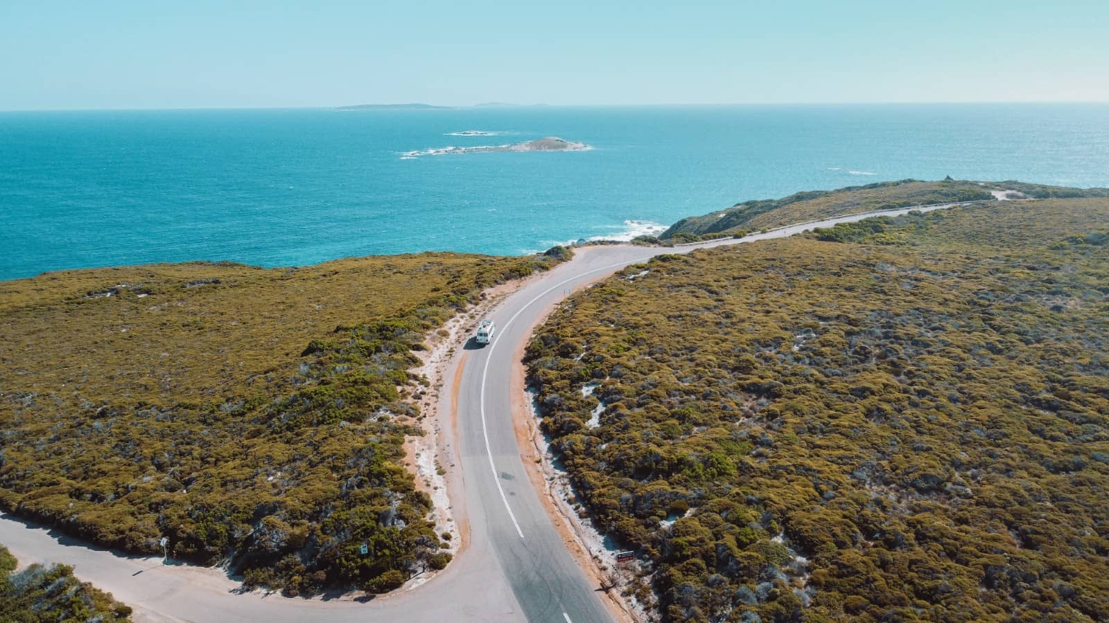 A winding coastal road curves along the rugged green shrubland of Esperance, Western Australia, with a white van driving toward the turquoise Indian Ocean. Small offshore islands and rocky outcrops dot the bright blue horizon under a clear sky. A must visit in any road trip from Adelaide to Perth