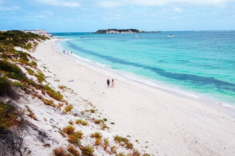 White sandy beach with turquoise water and people strolling along the shore in Hamelin Bay, an underrated coastal stop on a Melbourne to Perth road trip. Offshore boats and a small island are visible under a partly cloudy sky.