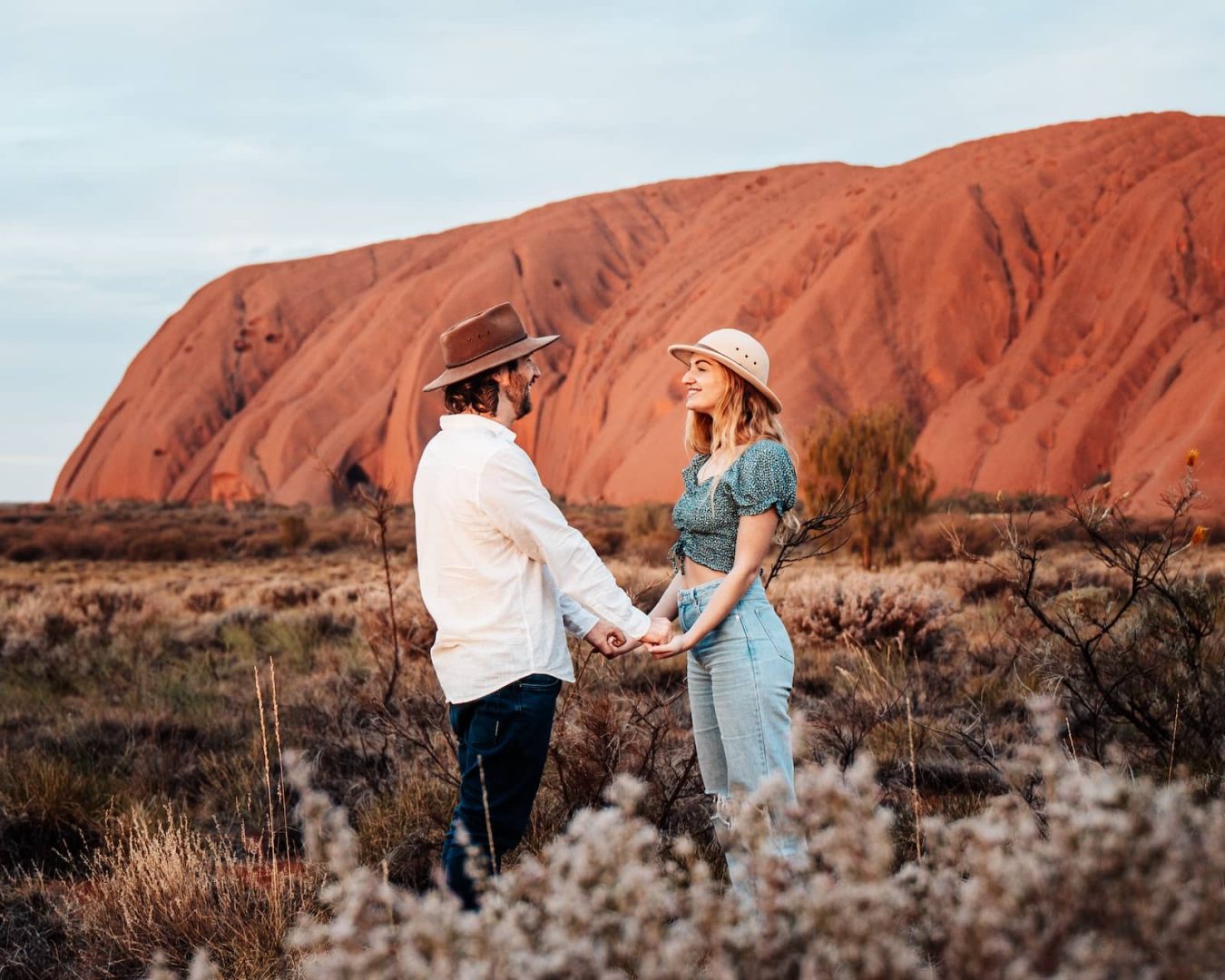 A couple stands holding hands and smiling at each other in front of the massive red rock of Uluru during an alice springs to uluru road trip. The dry desert shrubs and golden light highlight the iconic outback landscape.