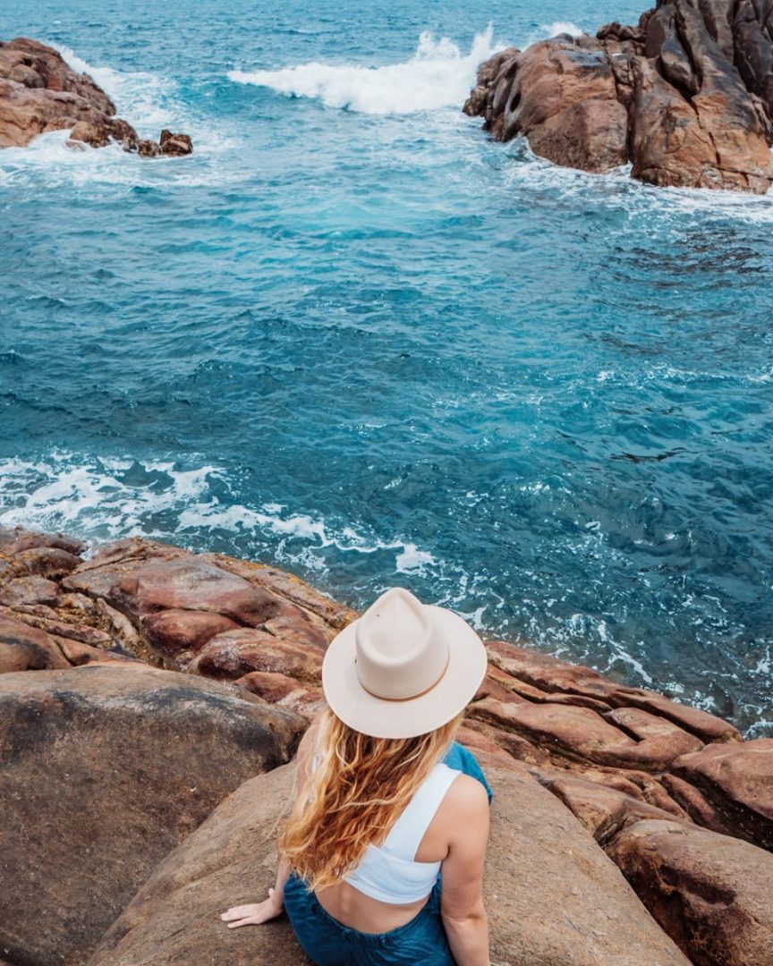 A woman in a wide-brim hat sits on coastal rocks, watching waves crash near Canal Rocks, Margaret River, on a sunny day. A popular scenic stop on a south west australia road trip 7 days adventure.