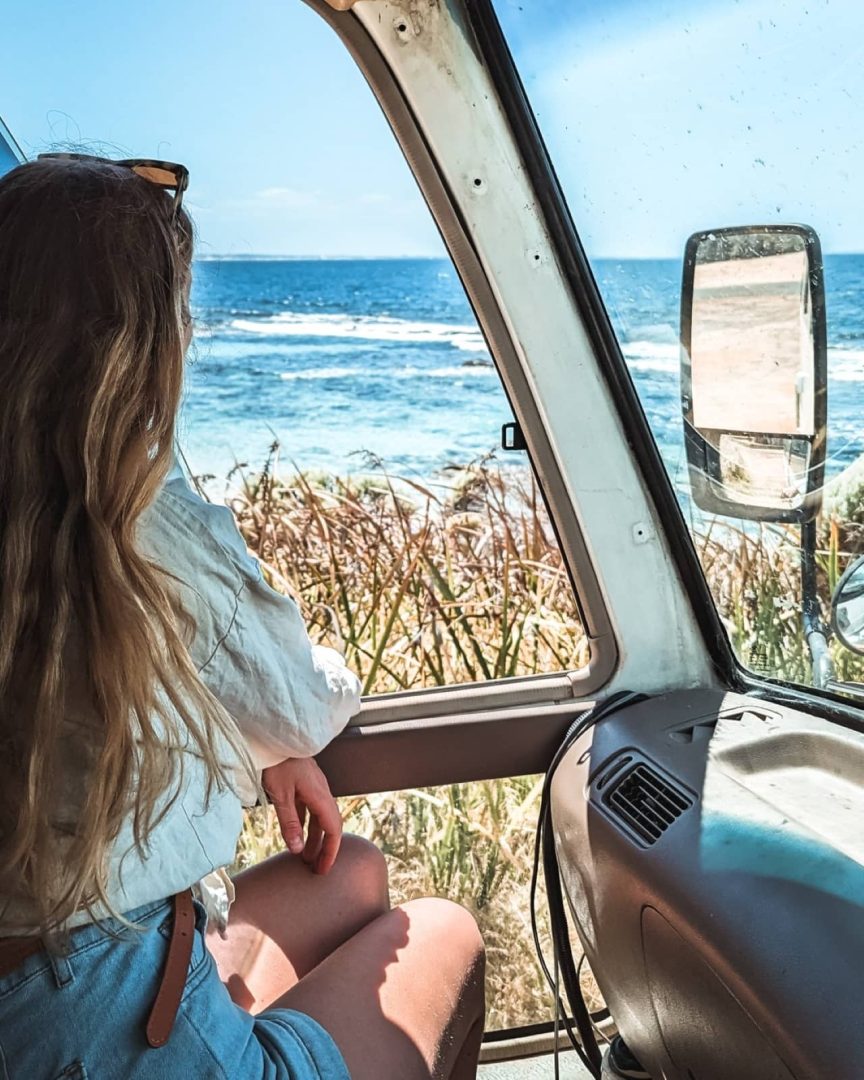 A woman sits in a campervan parked beside the coast, gazing out at Flinders Bay, Augusta, WA on a sunny day. Van life moments like this define the freedom of a south west australia road trip 7 days.