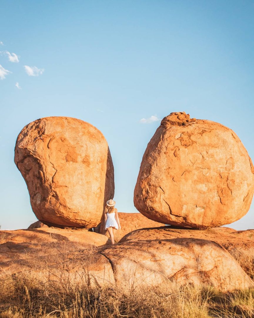 Woman in a white dress and hat stands between two massive rounded boulders at the Devils Marbles in the Northern Territory, a must-see on a Melbourne to Darwin road trip.