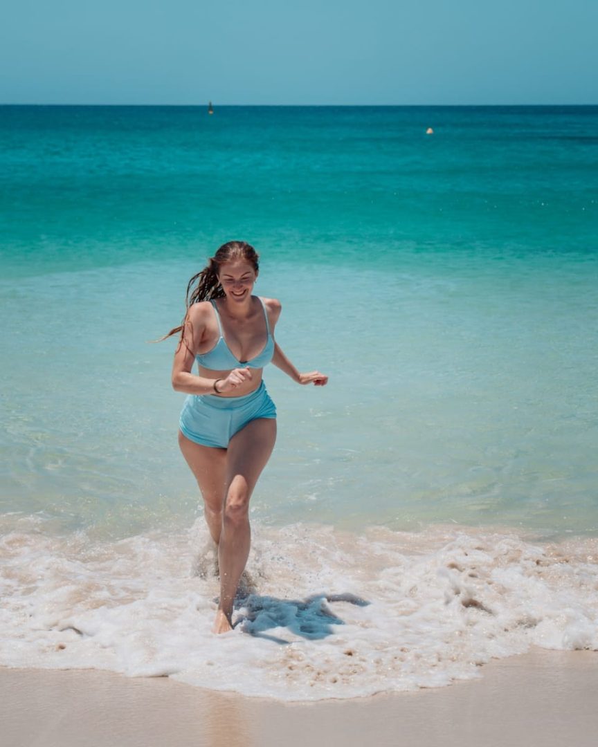 A smiling woman in a blue retro-style bikini runs from the ocean onto a white sandy beach in South West Australia. Beach swims like this are a daily joy on a south west australia road trip 7 days.