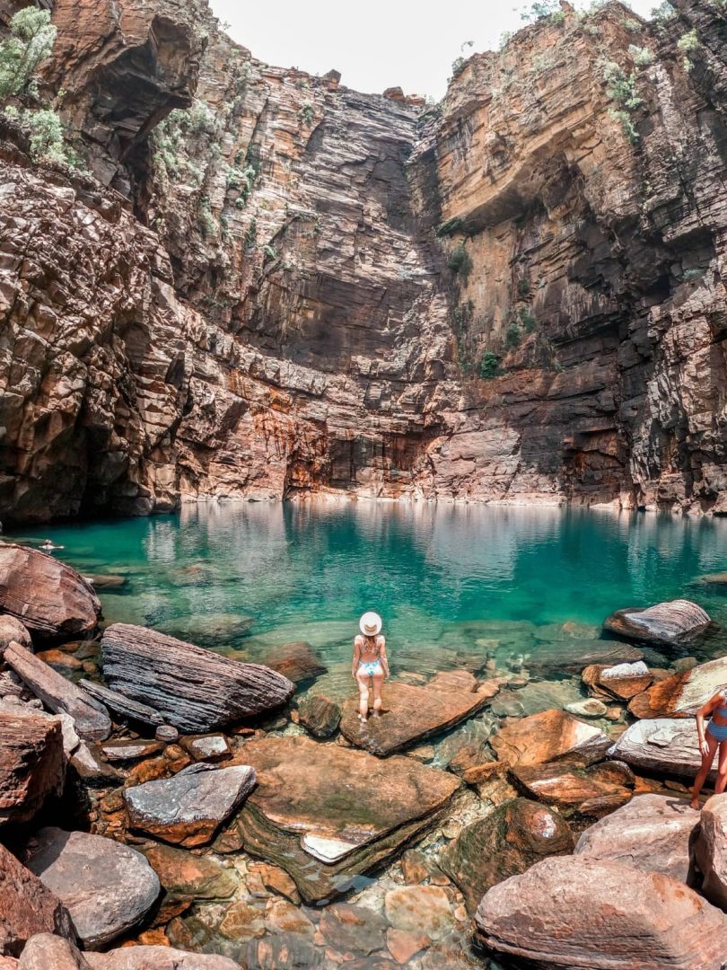 Woman sits on rocks beside the bright turquoise waters of Jim Jim Falls in Kakadu National Park, one of the top natural highlights of a Melbourne to Darwin road trip.