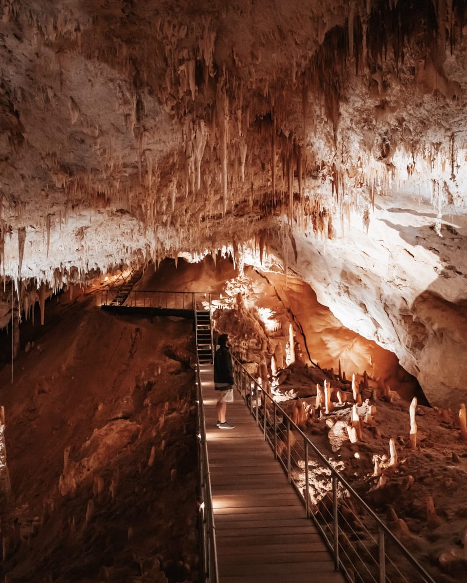 A person stands inside a limestone cave, surrounded by dramatic stalactites and stalagmites illuminated by warm lighting. Underground wonders like these feature on a south west australia road trip 7 days plan.