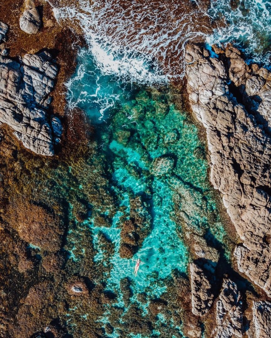 Aerial view of a woman floating in The Aquarium, Yallingup, a natural rock pool surrounded by vibrant turquoise water and jagged stone in South West Australia. Ideal for a scenic swim stop on a south west australia road trip 7 days.