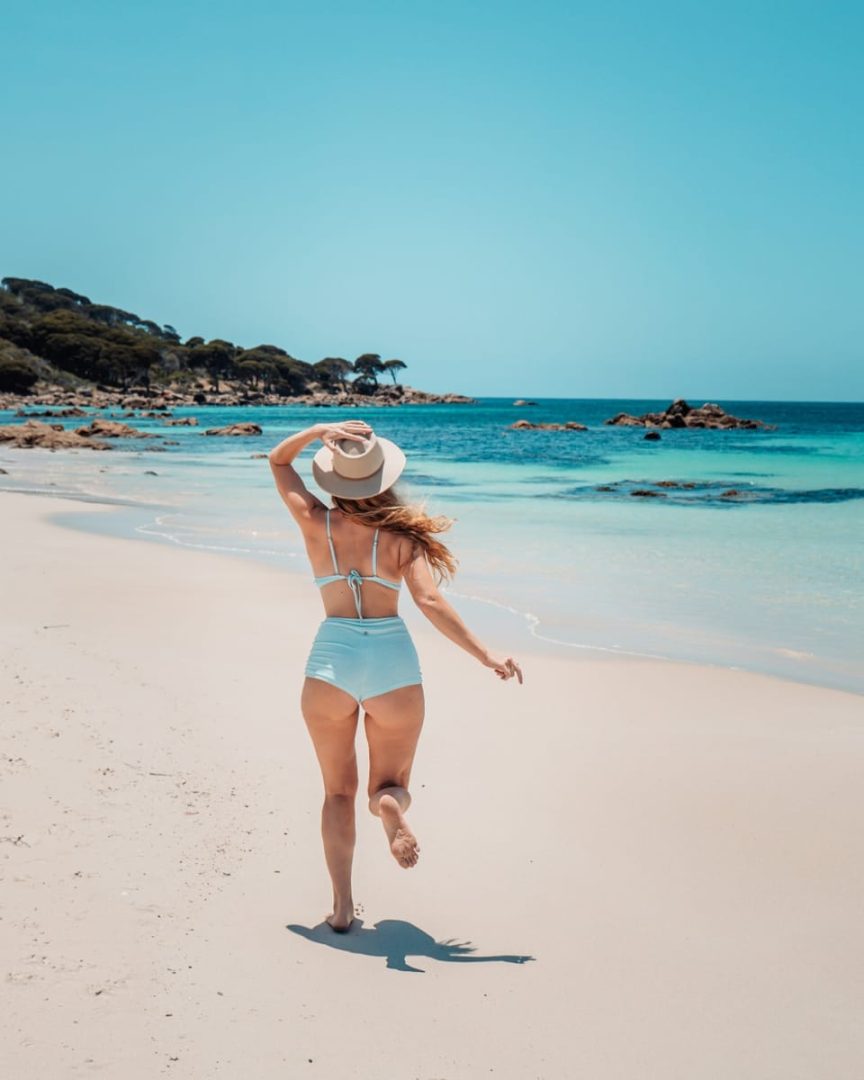 A woman in a retro-style blue swimsuit runs along a white sand beach with crystal-clear water in South West Australia. Beach-hopping like this is part of the charm on a south west australia road trip 7 days.