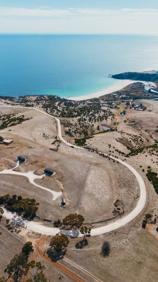 Birdโs-eye view of rolling dry hills and white sandy coastline with bright blue sea on Kangaroo Island, a detour possible during a Melbourne to Darwin road trip.