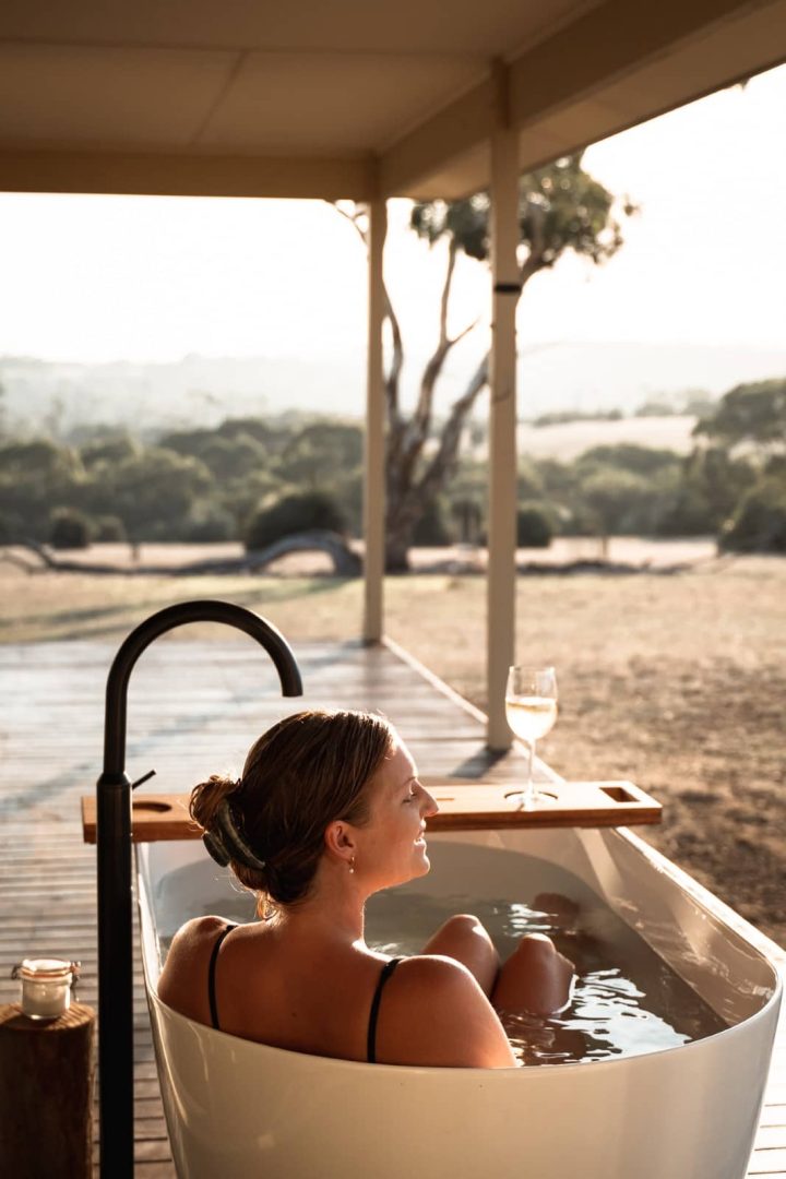 Woman soaking in an outdoor tub with a glass of white wine, overlooking scenic rolling hills on a kangaroo island road trip 3 days.