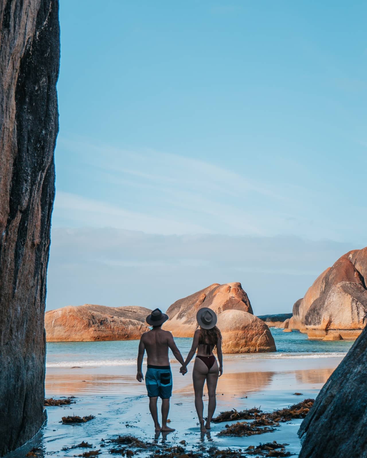 A couple walks hand in hand through a gap between massive granite boulders towards the ocean at Elephant Rocks, South West Australia. This photogenic stop belongs on every south west australia road trip 7 days plan.