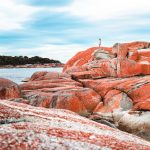A person standing atop striking red lichen-covered boulders at the Bay of Fires, a must-see stop on a 7 day Tasmania road trip.