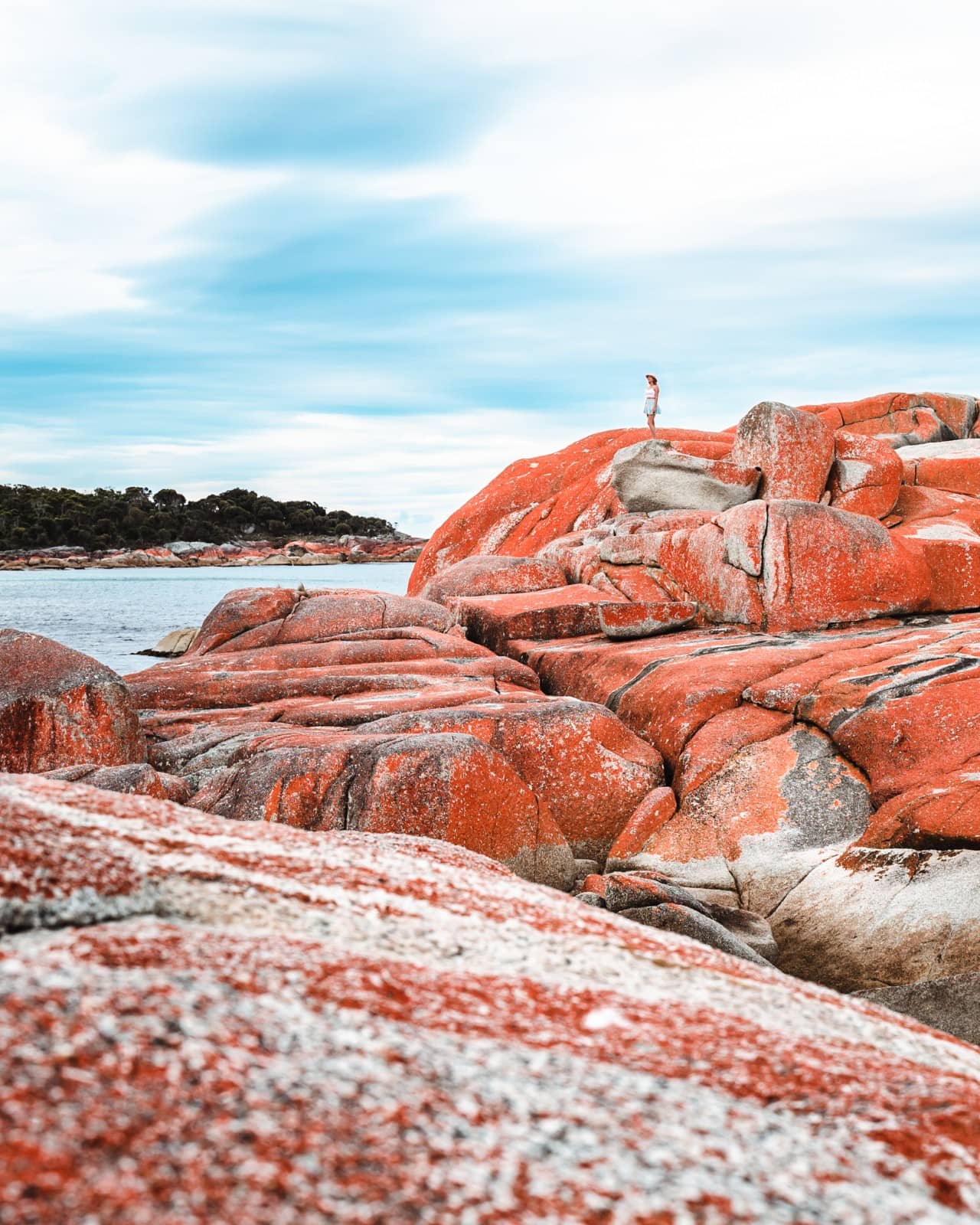 A person standing atop striking red lichen-covered boulders at the Bay of Fires, a must-see stop on a 7 day Tasmania road trip.