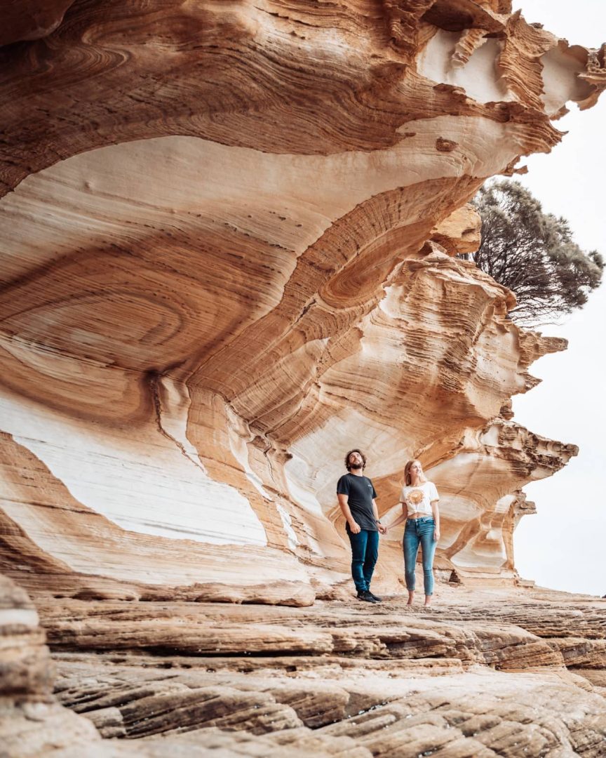A couple standing under swirling orange and white sandstone formations at the Painted Cliffs on Maria Island during a great eastern drive adventure.