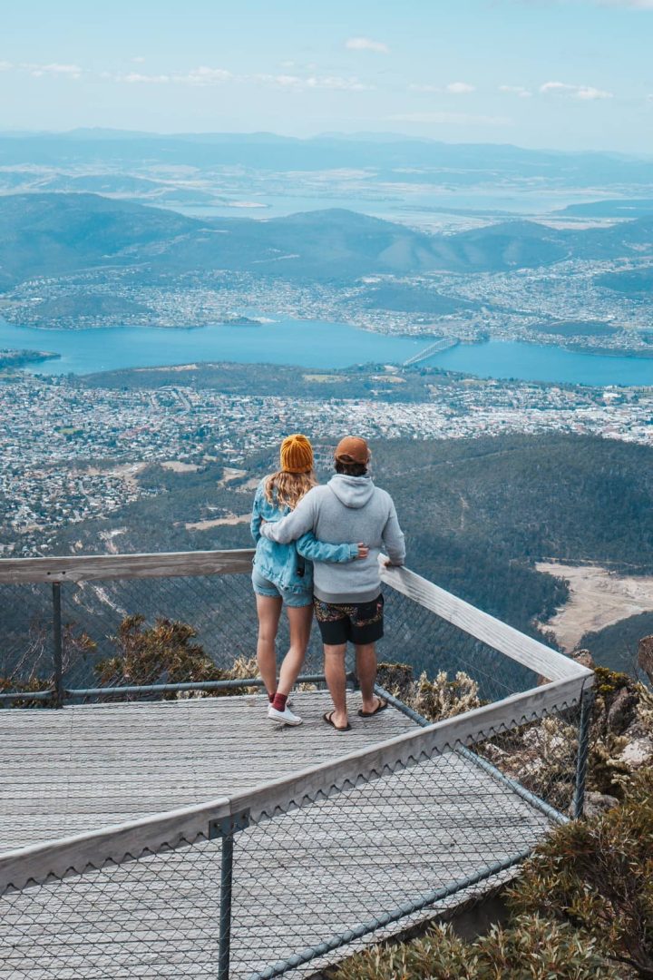 A couple stands at a lookout on Mount Wellington, gazing over Hobart and the Derwent River during a scenic 7 day Tasmania road trip.