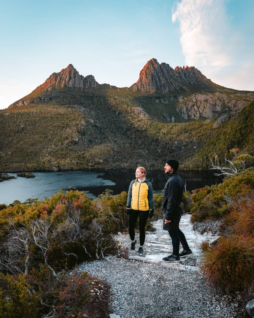 Two hikers on a gravel track overlooking Dove Lake with Cradle Mountainโs rugged peaks behind them at golden hour, a must-see stop when exploring things to do in Tasmaniaโs national parks.