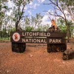 A smiling woman with both arms raised sits on a sign that reads "Litchfield National Park", marking her arrival at a major stop on a Darwin to Litchfield road trip.