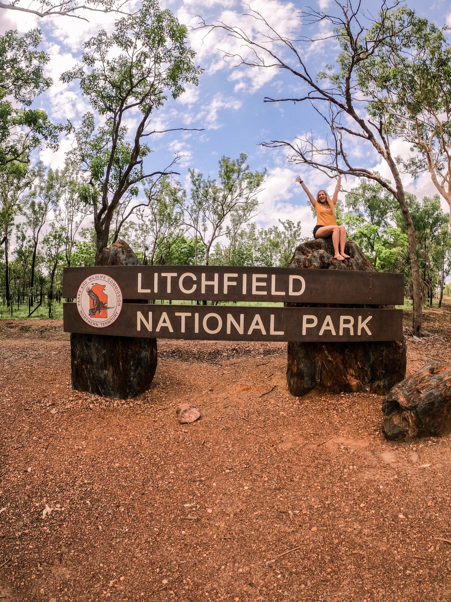 A smiling woman with both arms raised sits on a sign that reads "Litchfield National Park", marking her arrival at a major stop on a Darwin to Litchfield road trip.