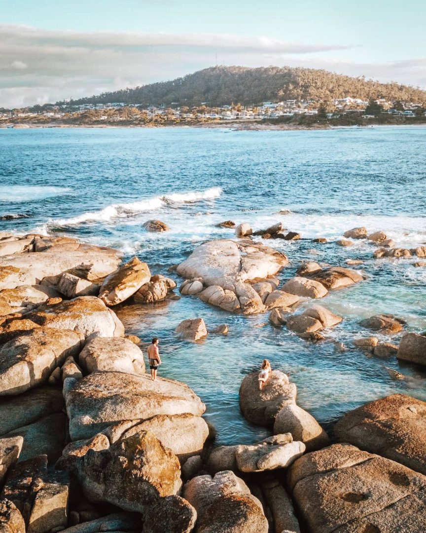 Two people sit and stand among large granite boulders beside clear blue water at Binalong Bay in the Bay of Fires. Waves wash through the rocks with the coastal town and wooded hills visible across the bay.