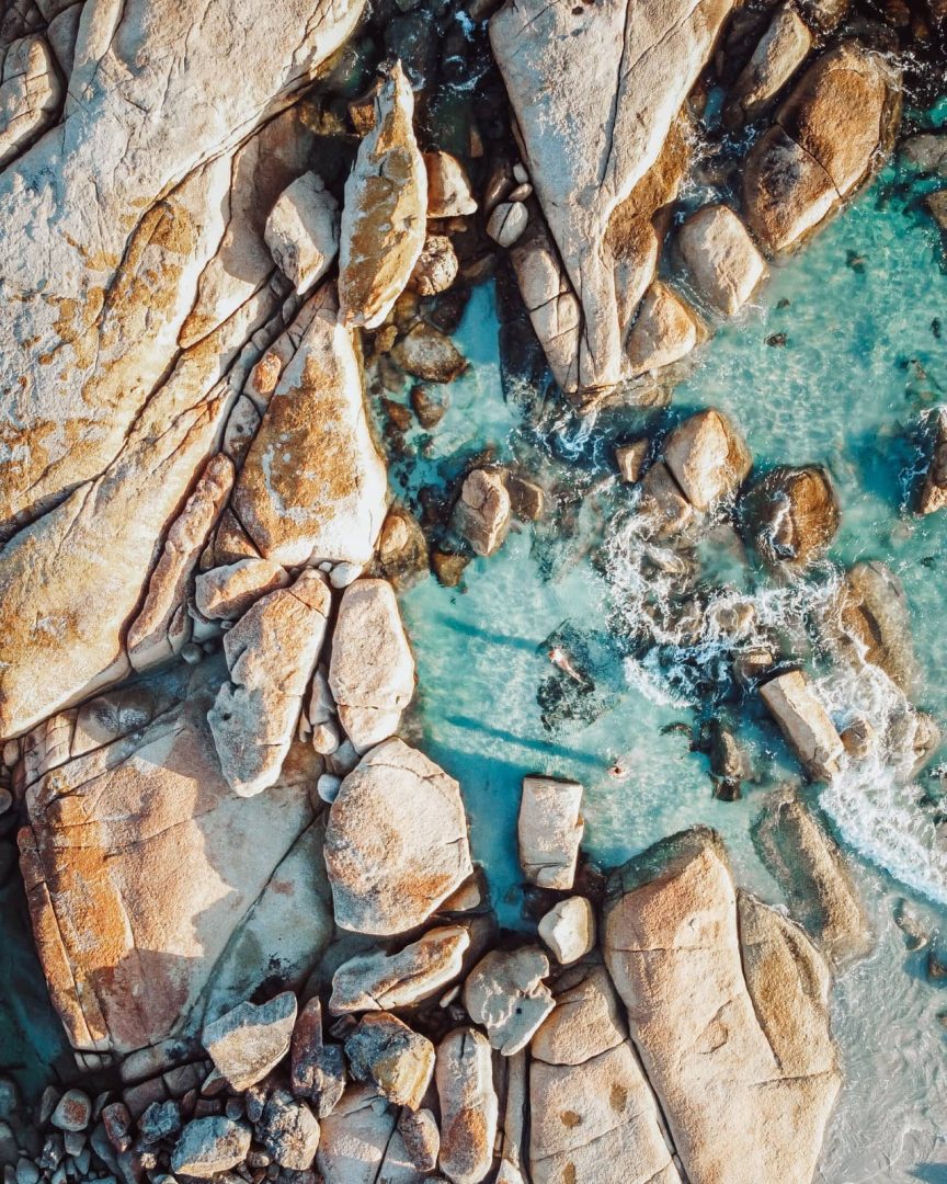 Aerial view of pale granite boulders surrounded by bright turquoise water at Binalong Bay in the Bay of Fires. Small waves break between the rocks along this coastal stop on a Tasmania road trip from Devonport.