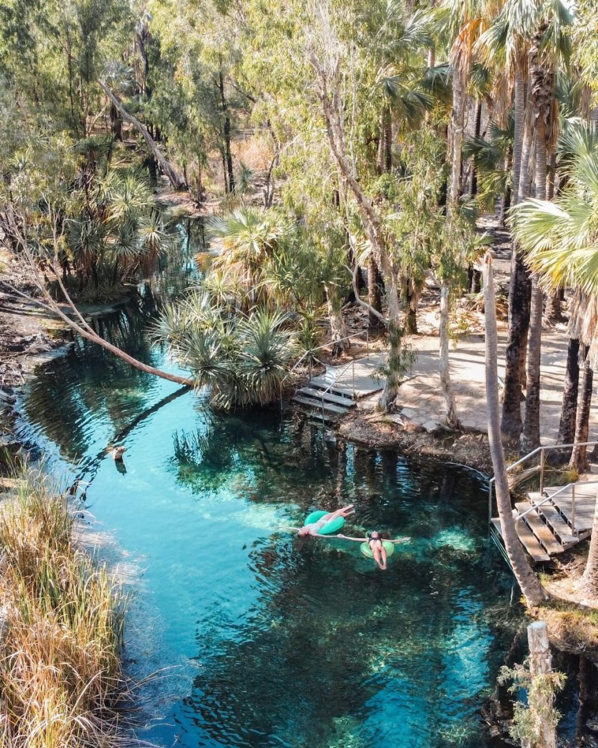 Two people drift downstream on pool noodles through the turquoise water of Bitter Springs. Palm trees and native vegetation line the narrow spring and make the float feel surrounded by nature.