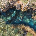 Two people float on pool noodles in the clear blue water of Bitter Springs beside a set of entry steps. Palms surround the natural swimming hole and show how secluded the spring feels.