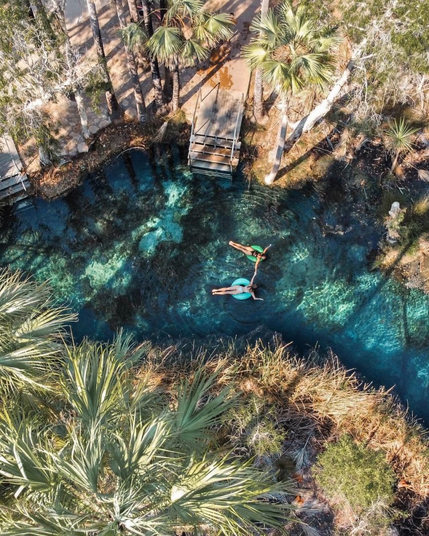 Two people float on pool noodles in the clear blue water of Bitter Springs beside a set of entry steps. Palms surround the natural swimming hole and show how secluded the spring feels.