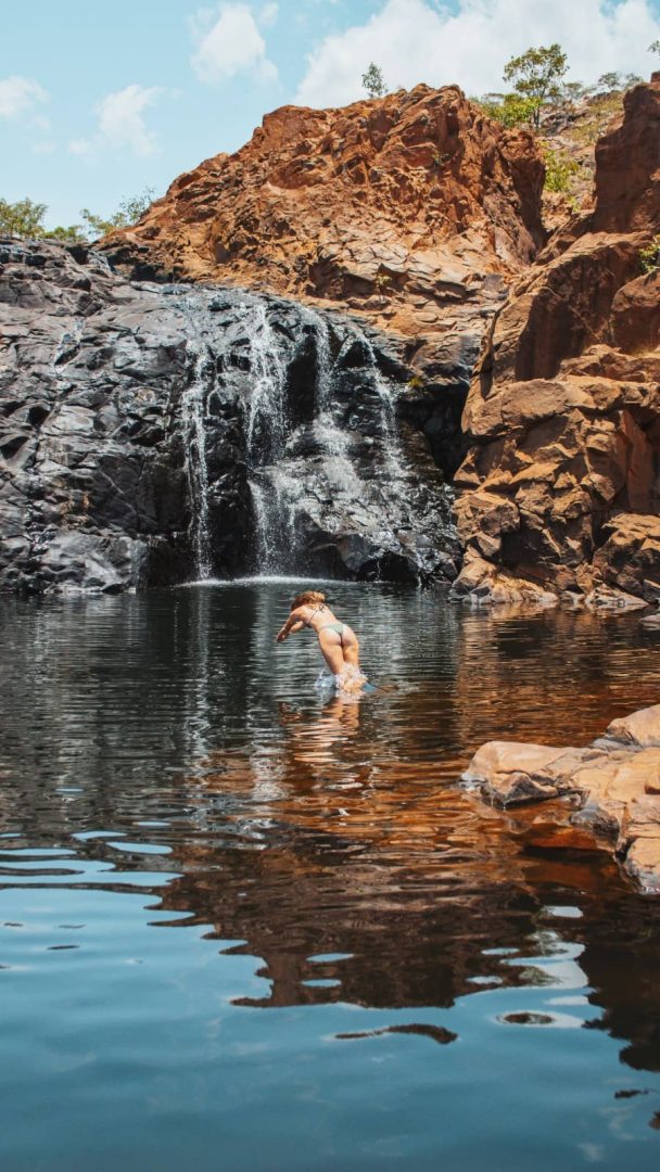 A woman dives into the natural pool beneath a waterfall at Leliyn Edith Falls. Red rocky cliffs frame the swimming spot visited on the return leg of the Darwin to Katherine weekend road trip.