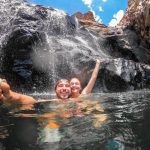 Two people swim beneath a rocky waterfall at Edith Falls in Nitmiluk National Park. Water cascades down the dark rock face behind them beneath a bright blue sky.