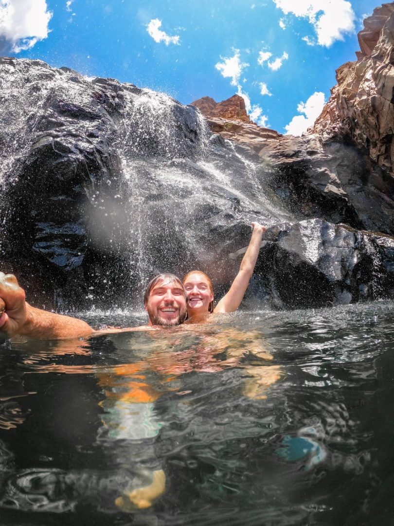 Two people swim beneath a rocky waterfall at Edith Falls in Nitmiluk National Park. Water cascades down the dark rock face behind them beneath a bright blue sky.