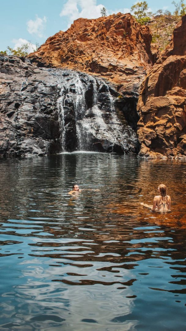 Two people swim in a dark natural pool below a waterfall at Leliyn Edith Falls. Orange cliffs surround the swimming hole in the northern section of Nitmiluk National Park.