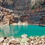 Turquoise plunge pool beneath the towering rock cliffs of Jim Jim Falls in Kakadu National Park. A scenic stop on a Darwin to Kakadu weekend road trip with visitors resting on rocks near the water.