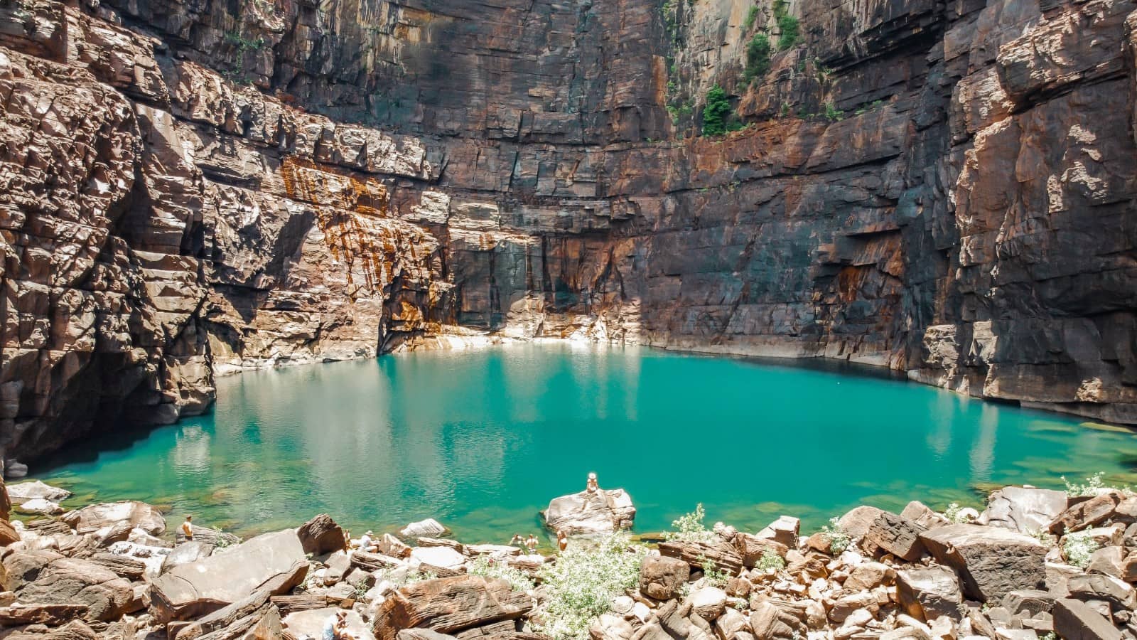 Turquoise plunge pool beneath the towering rock cliffs of Jim Jim Falls in Kakadu National Park. A scenic stop on a Darwin to Kakadu weekend road trip with visitors resting on rocks near the water.